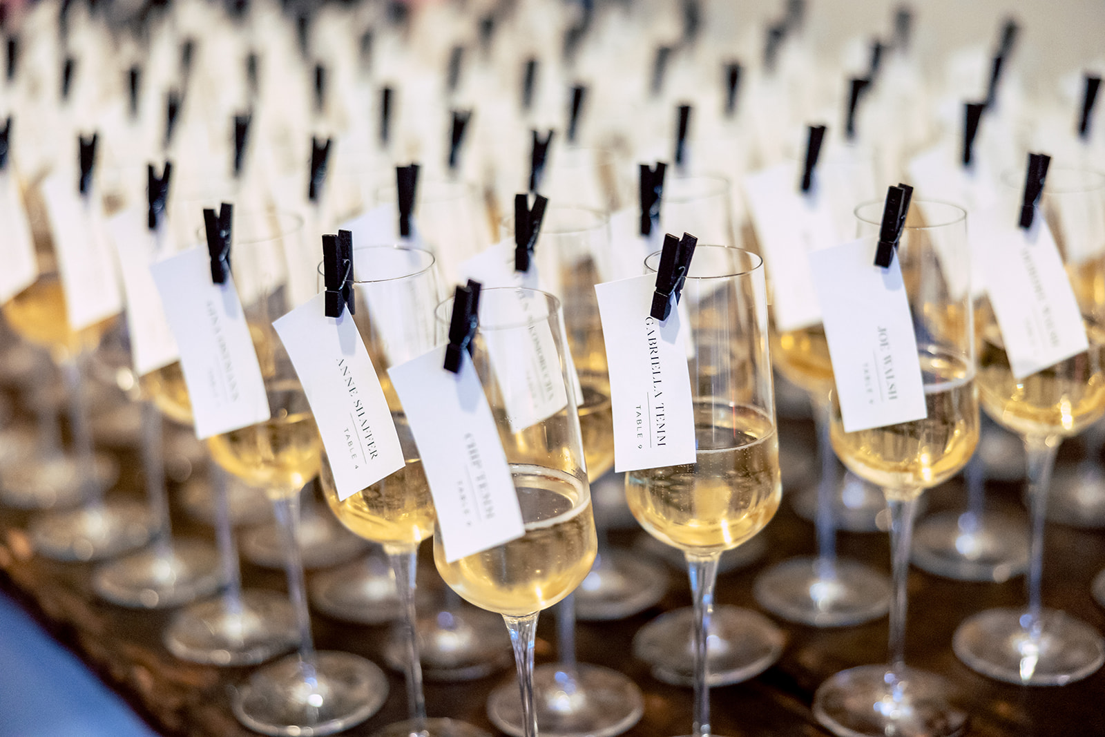 Rows of wine glasses filled with white wine, each with a small card clipped to the rim using black clothespins. The glasses are arranged neatly on a wooden surface.