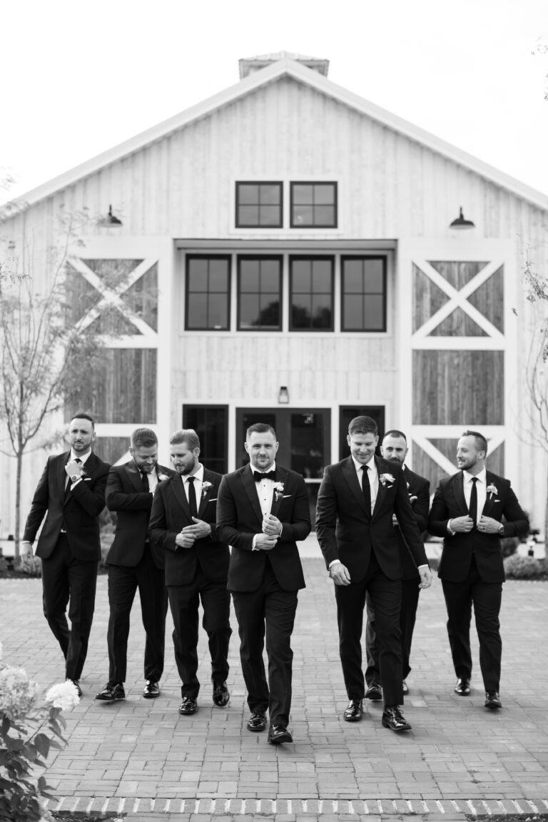 Seven men in suits walk confidently outside a rustic barn venue. They are all in black suits with boutonnières, with the central figure in a tuxedo. The barn has large doors and windows, and the surroundings are landscaped with trees and plants.