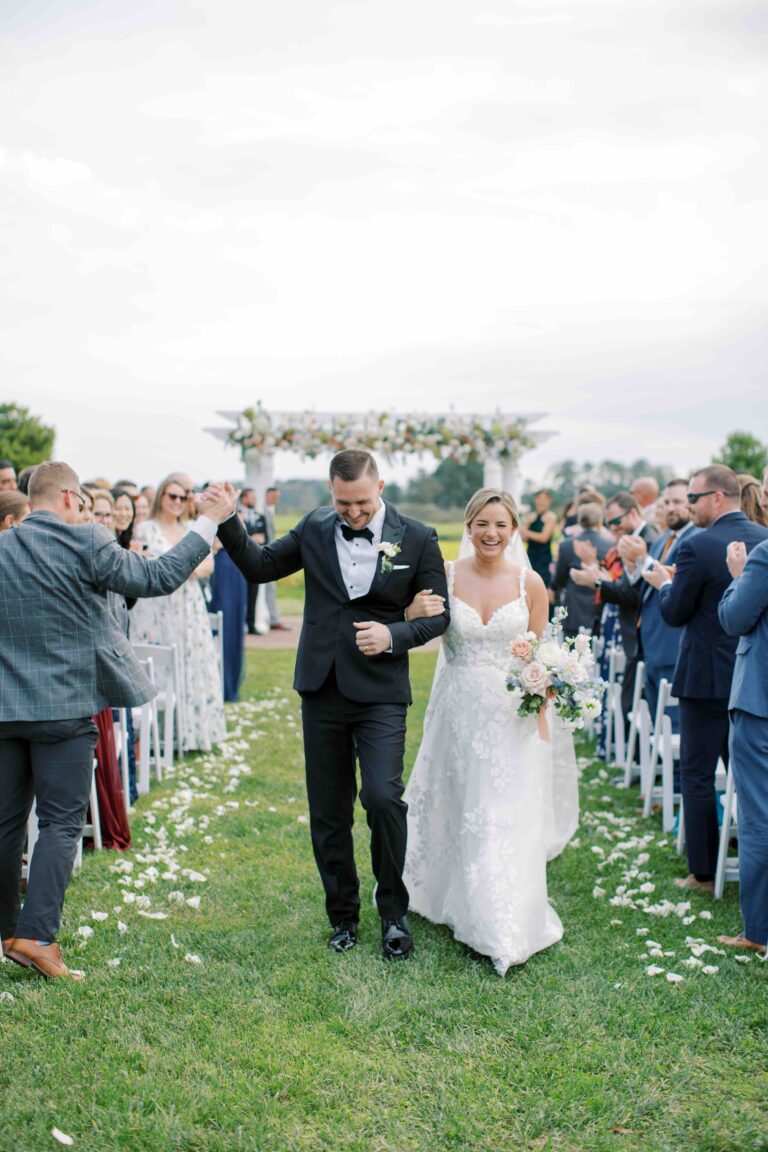 A bride and groom walk arm-in-arm down a grassy aisle outdoors, smiling as guests clap and celebrate. The bride wears a white dress and holds a bouquet; the groom is in a black tuxedo. White petals line the aisle.