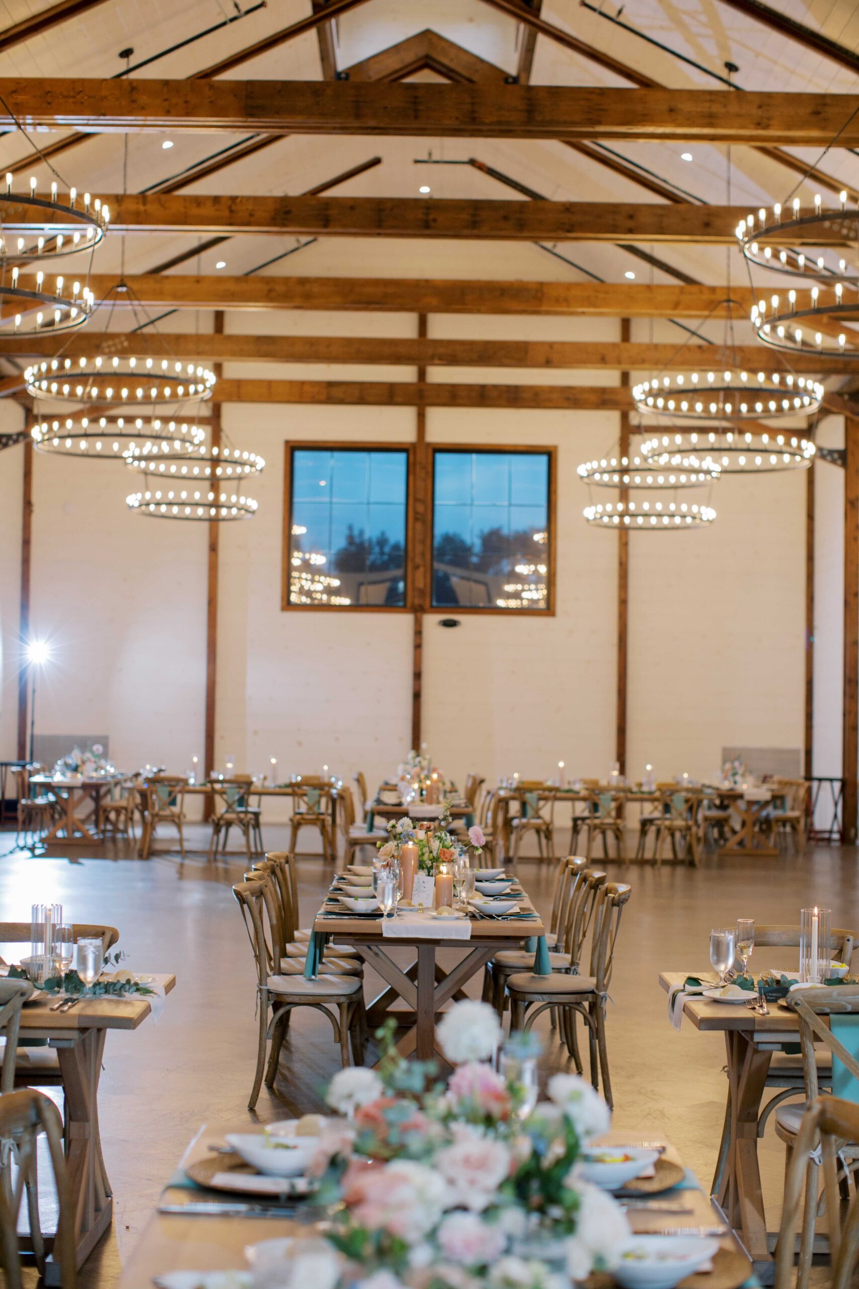 Spacious, elegantly decorated wedding venue with long wooden tables adorned with floral centerpieces and place settings. Large, circular chandeliers hang from a high, wooden-beam ceiling. A window is visible in the background.