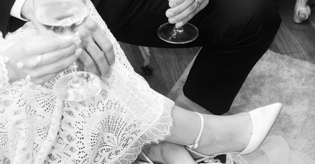 Black and white photo of a seated couple, possibly at a wedding. The woman wears a lace dress and white heels, and both hold wine glasses. A veil drapes across the floor.