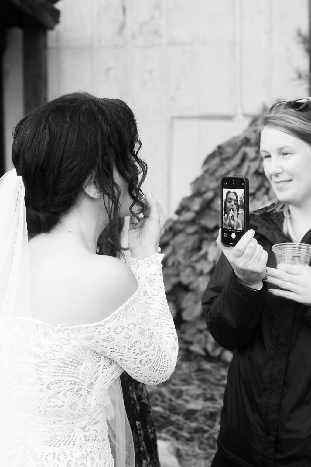 A bride in a lace dress and veil smiles while looking at herself in a phone held by another woman, who is holding a drink. The scene is outdoors, and the image is in black and white.