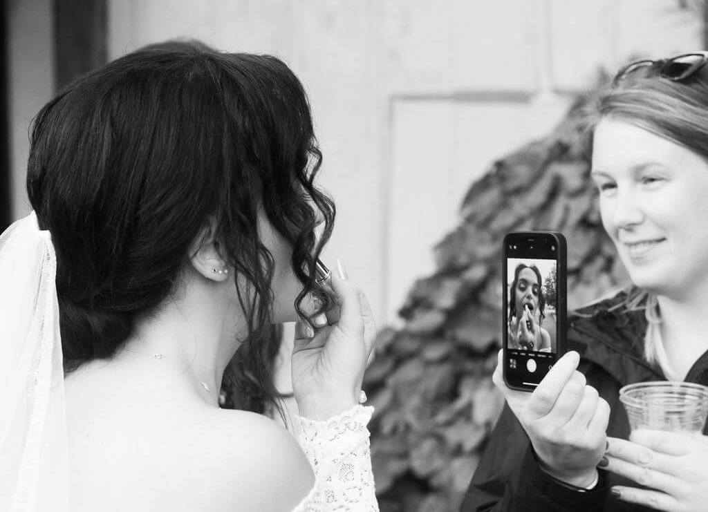 A bride holds lipstick near her lips, adjusting her makeup. Another person captures the moment on a smartphone. The scene is casual and candid, highlighting a joyful pre-ceremony preparation. The photo is in black and white.