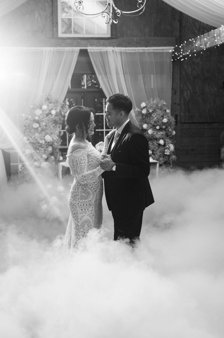 A bride and groom share a romantic first dance surrounded by low-lying fog in an elegant, flower-decorated venue, with soft sunlight streaming through a window.