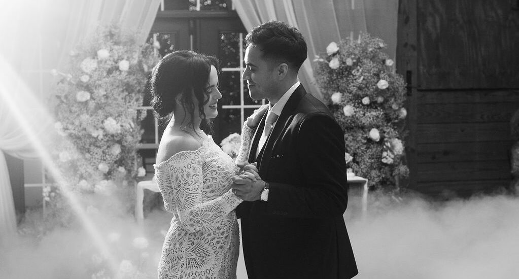 A bride and groom share a romantic first dance surrounded by mist in an elegantly decorated venue with flowers and draped curtains. Sunlight streams in, creating a dreamy, magical atmosphere.