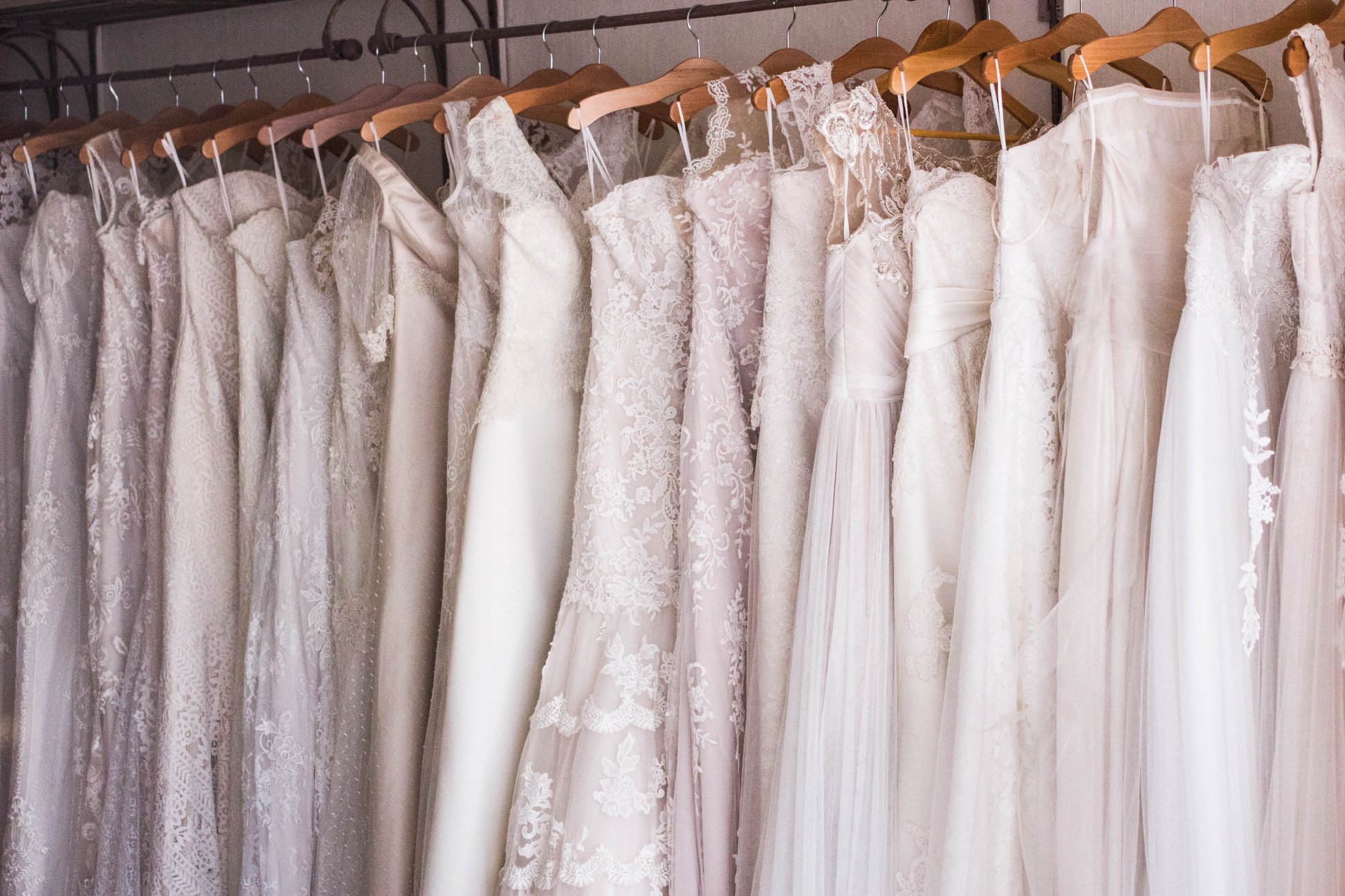 A row of white and ivory wedding dresses with lace and tulle details hang on wooden hangers in a bridal shop.