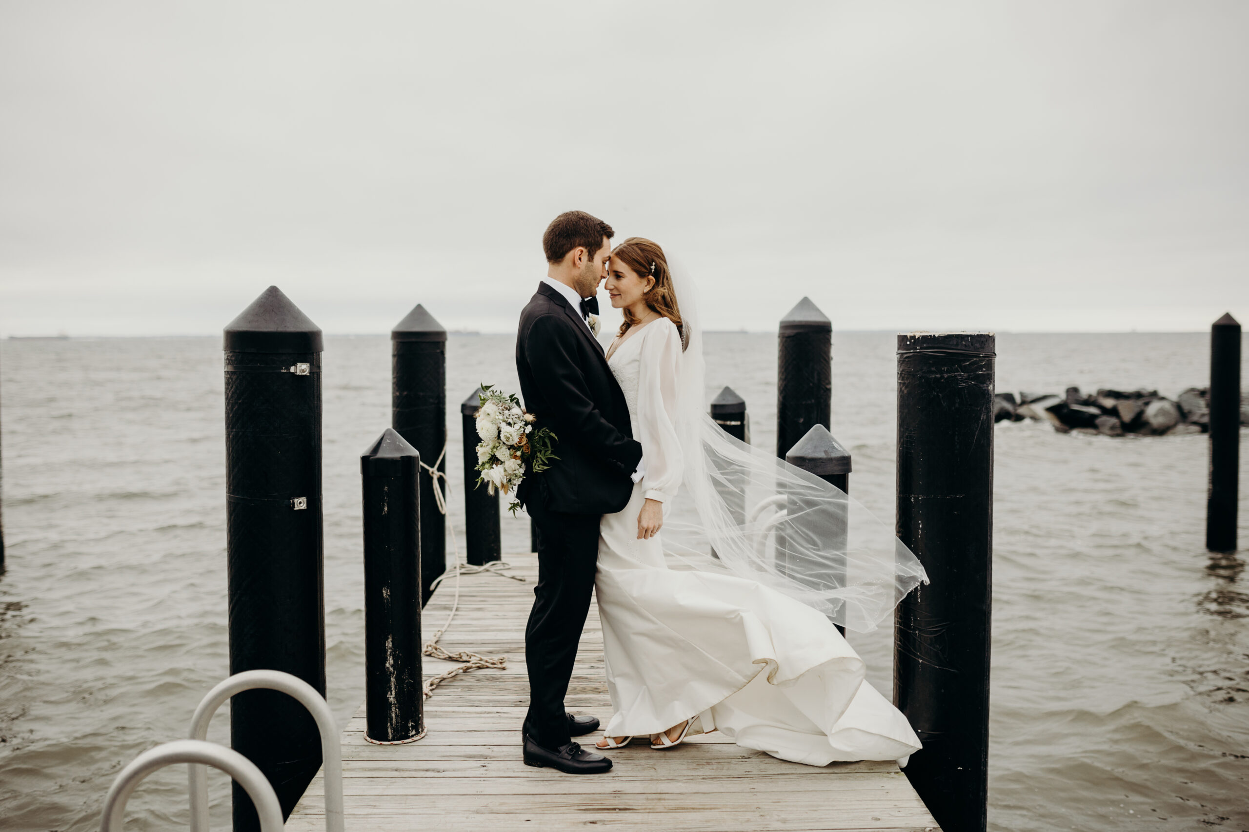 A bride and groom stand on a wooden dock surrounded by water. The groom, in a black suit, holds a bouquet. The bride, in a white dress with a long veil, smiles at him. Several black posts line the dock, and the sky is overcast.