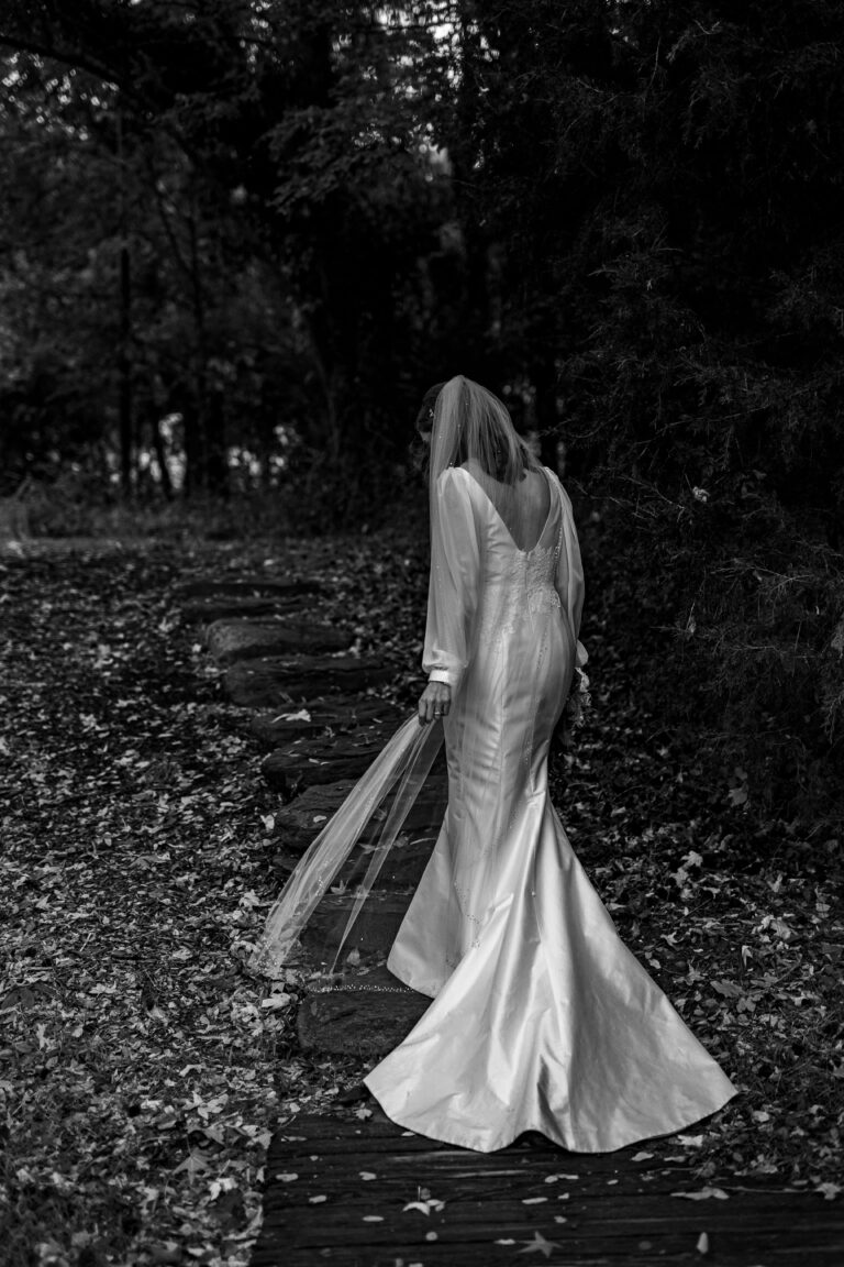 A bride in a flowing gown and veil walks along a rustic wooden path surrounded by foliage. Fallen leaves cover the path, and trees line the area, creating a serene, woodland atmosphere. The image is in black and white.