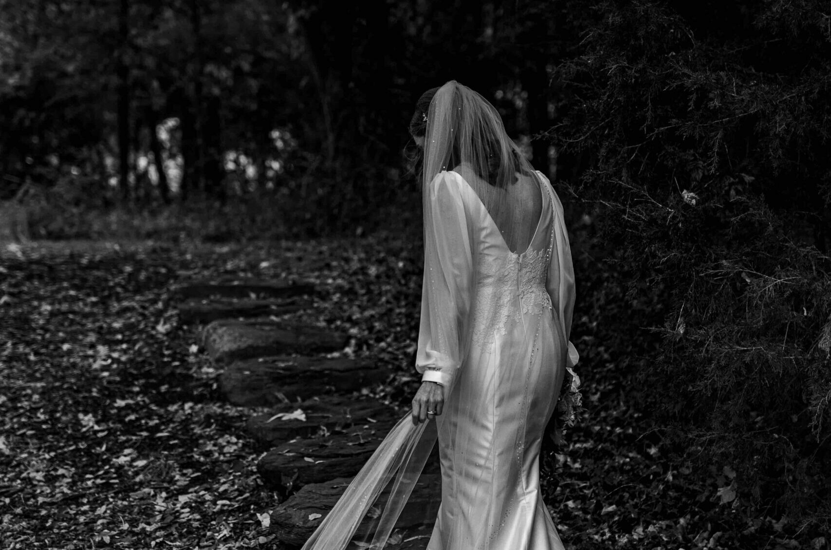 A bride in a long-sleeved wedding dress and veil walks alone on a leaf-strewn path in a wooded area, facing away from the camera. The image is in black and white.
