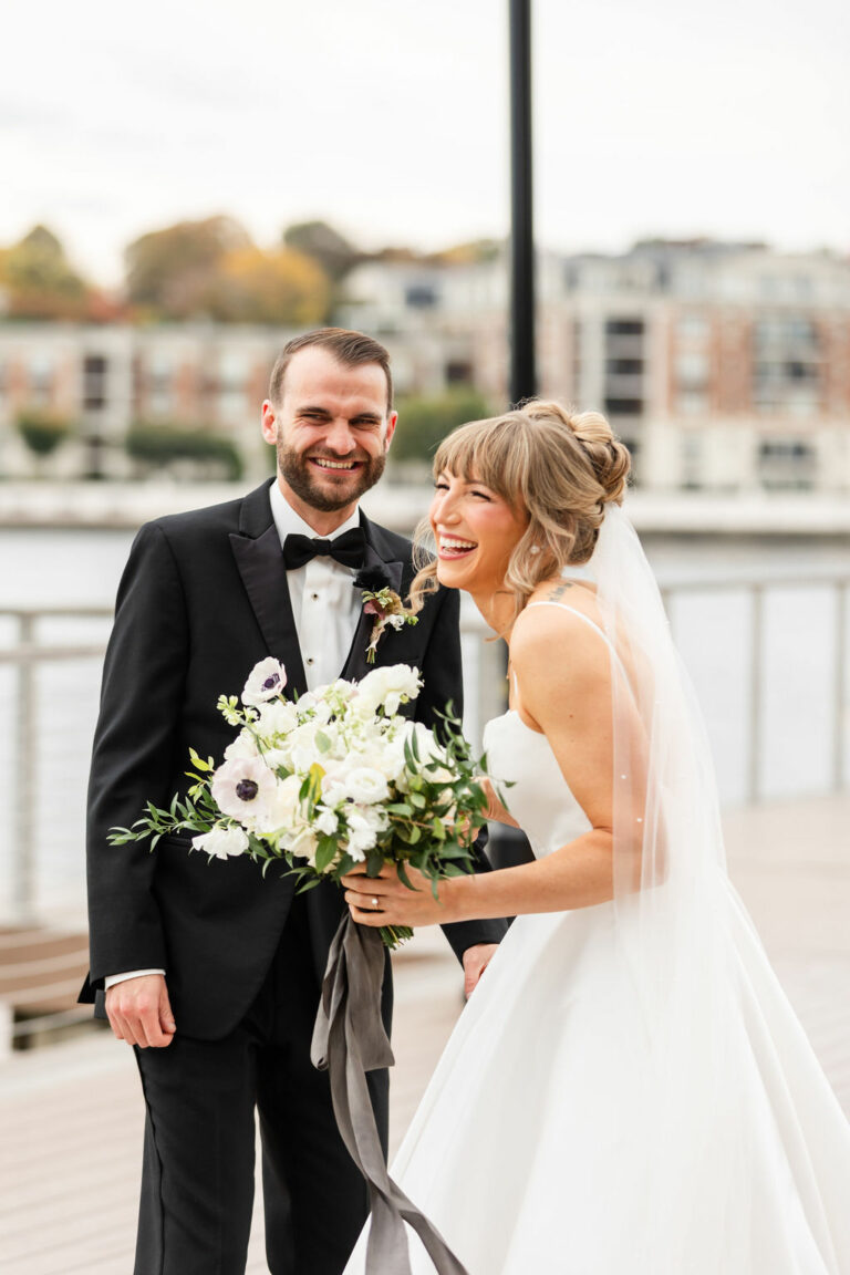 A bride in a white dress and veil holds a bouquet of white flowers and laughs joyfully next to a groom in a black tuxedo. They stand on a boardwalk with blurred buildings and a river in the background.