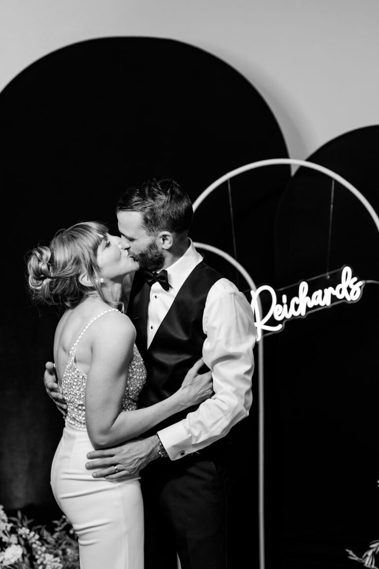 A couple sharing a kiss at a wedding reception. The woman is in a sparkling sleeveless dress, and the man is in a tuxedo. A neon sign in the background displays the word 