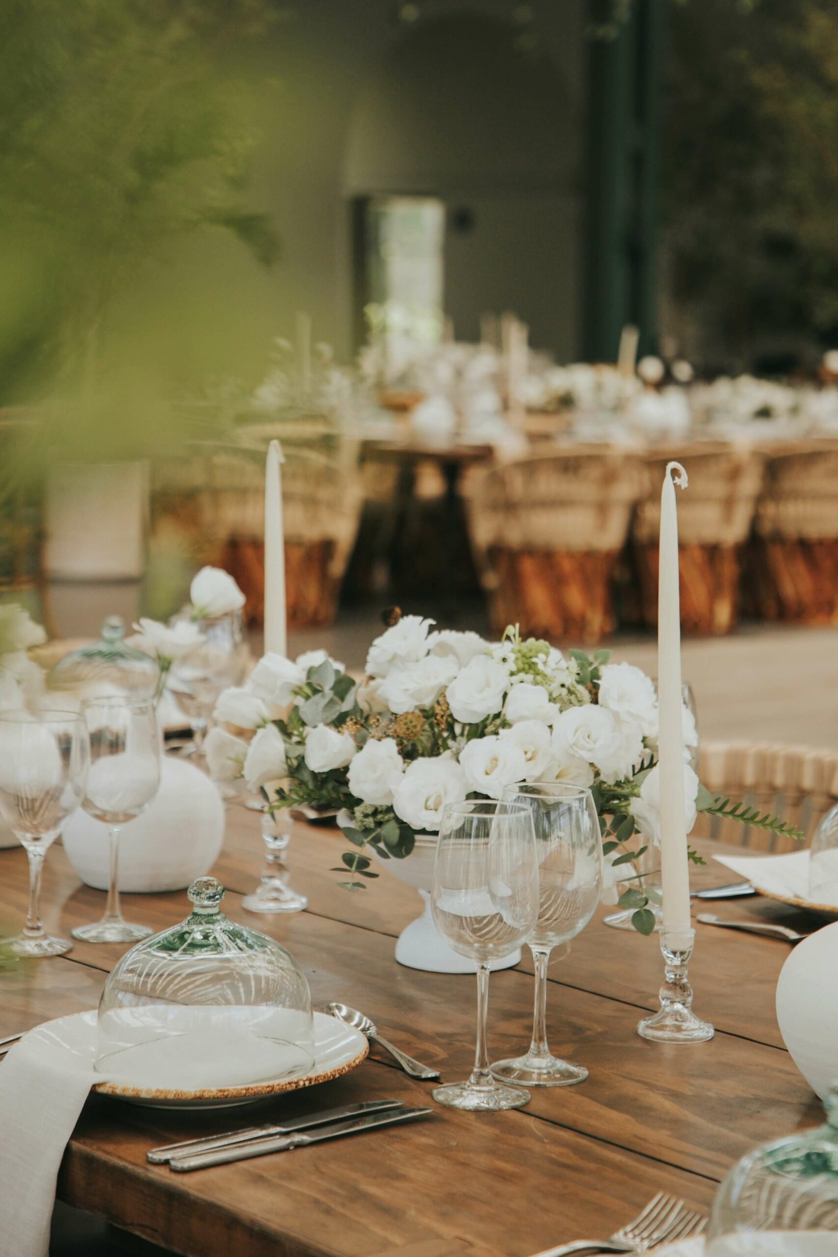 Elegant outdoor dining setup with a wooden table featuring white roses in a vase, tall white candles, glass plates with cloches, and wine glasses. In the background are more tables with similar arrangements, set in a lush garden setting.
