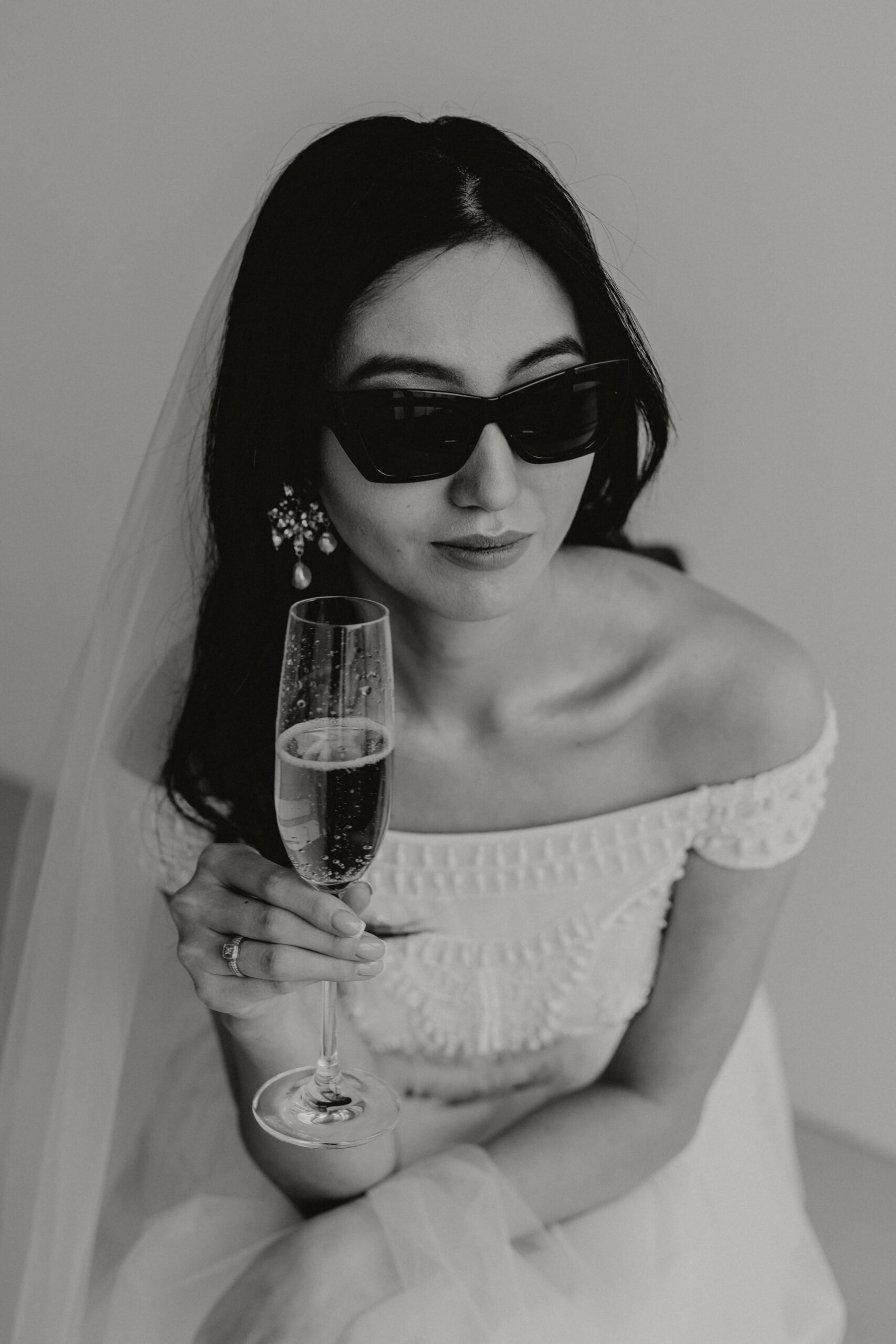 A woman in a white wedding dress and veil holds a champagne glass. She wears dark sunglasses and has long, dark hair. The dress features an off-shoulder design with lace details. The image is in black and white.