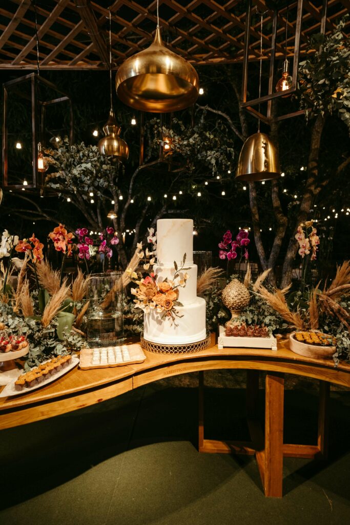 A two-tiered white cake with orange and red flowers sits on a wooden table. Surrounding the cake are various pastries and desserts. The table is decorated with greenery and dried pampas grass. Background includes hanging golden lights and festive decor.