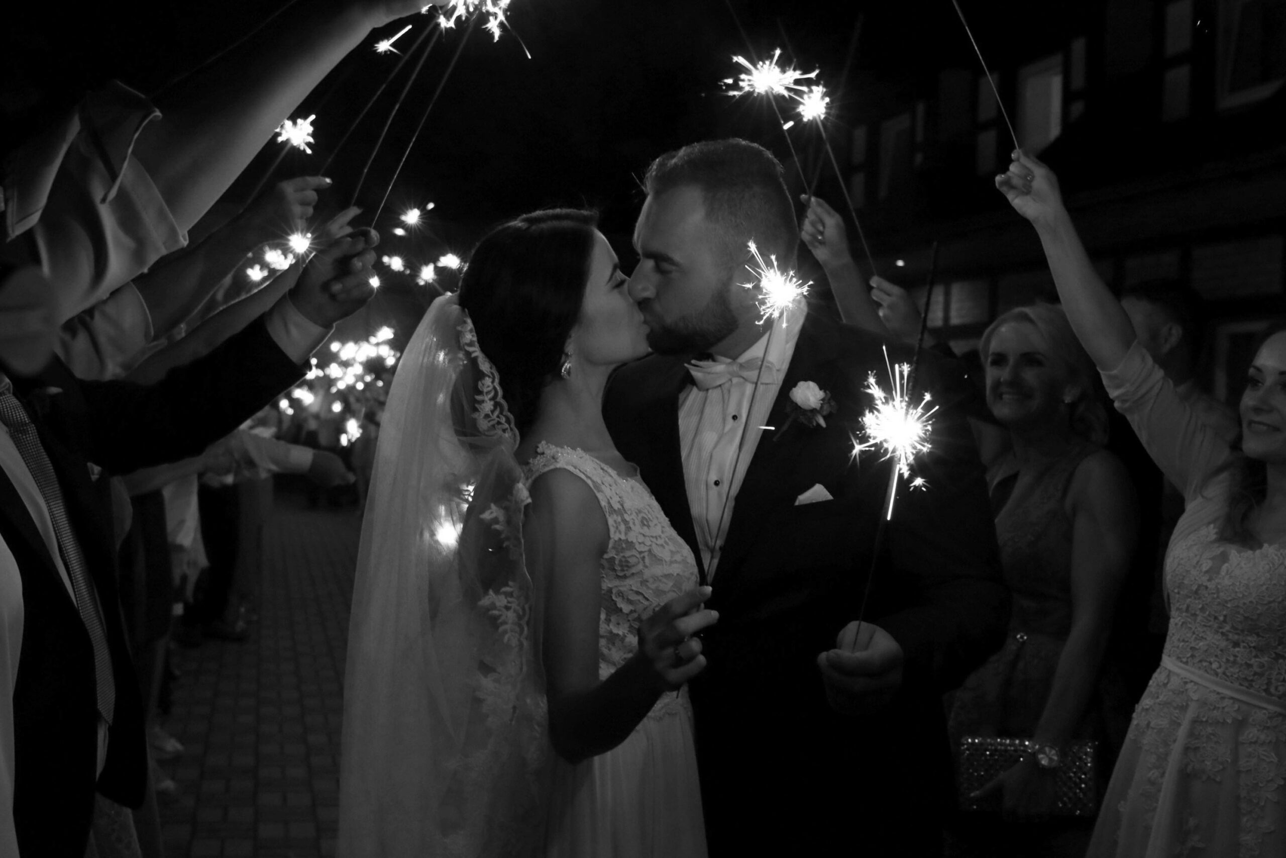 A bride and groom kiss under a canopy of sparklers held by guests at night. The bride wears a veil and a lace dress, and the groom is in a suit with a bow tie. Guests smile and celebrate around them in a joyful atmosphere.