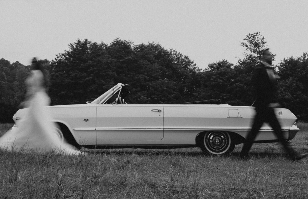 A black and white photo shows a classic convertible car parked on grass. A bride and groom, blurred in motion, walk past on opposite sides of the car. Trees fill the background.