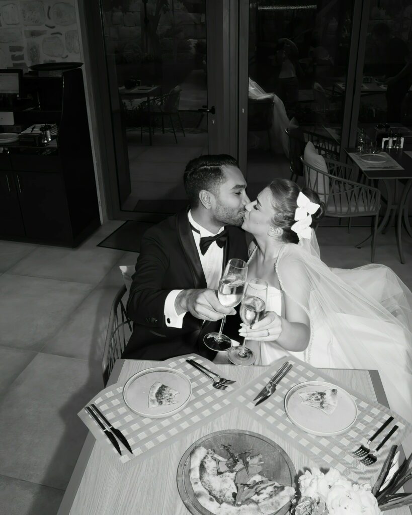 A bride and groom share a kiss while seated at a table, toasting with champagne glasses. The table is set with plates of food, including a pizza. They appear happy and are dressed in wedding attire.